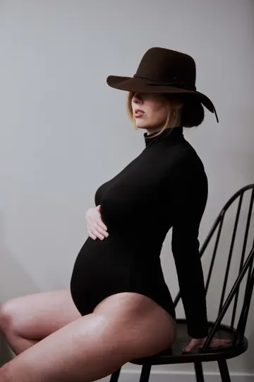 Pregnant woman in black bodysuit and brown hat, seated on black chair against neutral wall.