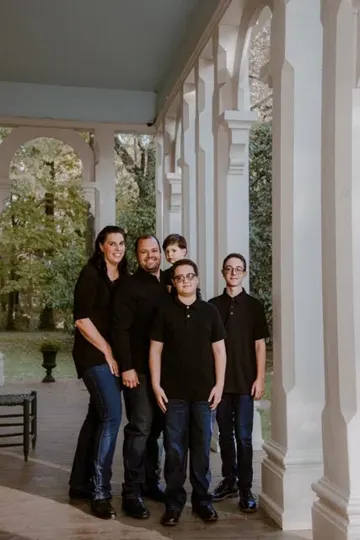 A family stands on a porch with white columns, sunlight filtering through trees.