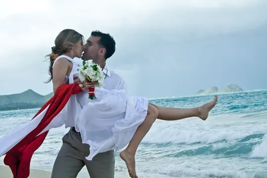A couple embraces on a beach, with ocean waves and distant hills under a cloudy sky.