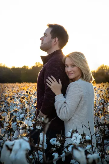 A couple embraces in a cotton field at golden hour, bathed in warm light.