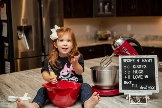Young child sits on kitchen counter, mixing in red bowl, with blackboard sign.