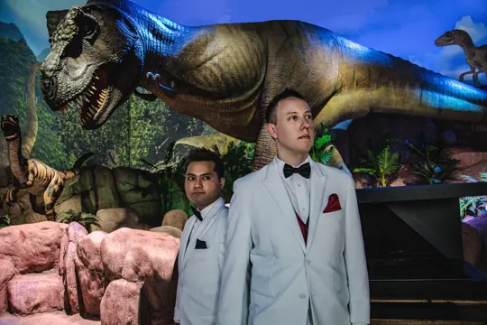 Two men in white suits stand before a life-sized T-Rex exhibit under blue lighting.