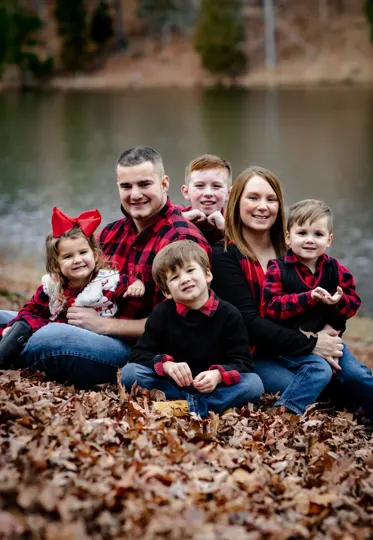 A family sits on fallen leaves by a calm lake, autumn light softening their coordinated red plaid and black attire.