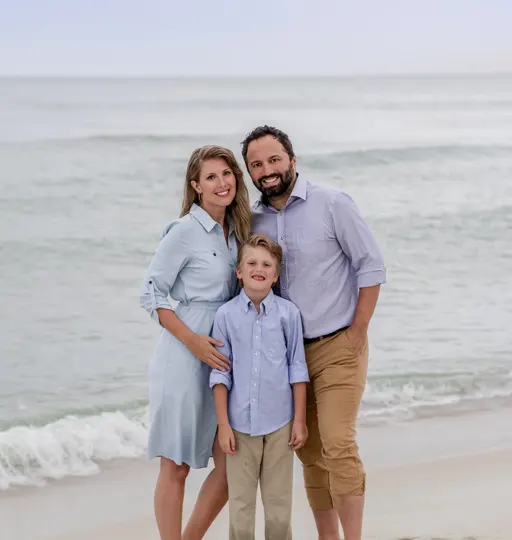 A family stands on a sandy beach, framed by gentle ocean waves and soft, diffused light.