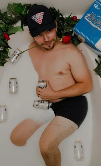 A person reclines in a bathtub, surrounded by cans and a box, with a rose in their mouth.