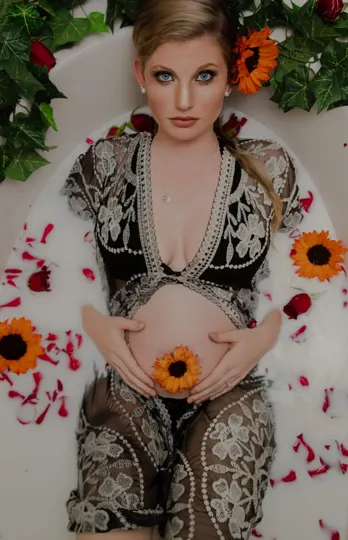 Pregnant woman in lace-patterned black dress, sunflowers, and rose petals in a tub.