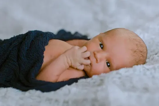 A newborn rests on a soft, light gray blanket, wrapped in dark blue fabric.